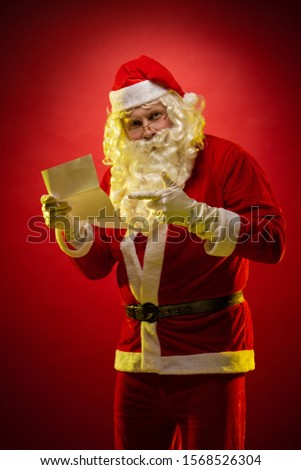 Male actor dressed as Santa Claus holds a sheet of paper in his hands a Christmas letter reads and poses on a dark red background