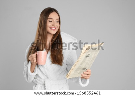 Beautiful young woman in bathrobe reading newspaper on light grey background