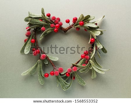 Decorative mistletoe wreath with frosted leaves and red berries shaped as a heart from above. Flat lay, top view on trendy metallic green paper. 