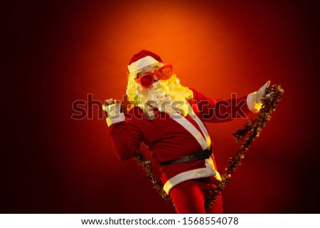 Male actor in a costume of Santa Claus in large pink glasses and with a red garland of tinsel in his hands dancing and posing on a dark red background