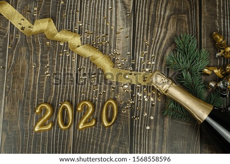 Champagne bottle with golden tinsel, ribbon and confetti on a dark wooden background. Golden bow, Christmas tree branch, bottle of champagne and numbers 2020 on a textured dark background. Flat lay