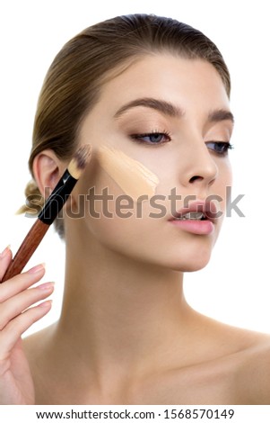 Young woman applying facial foundation cream at her face. Beauty model with perfect fresh skin and long eyelashes cares about her skin at home. Makeup Concept. Royalty-Free Stock Photo #1568570149