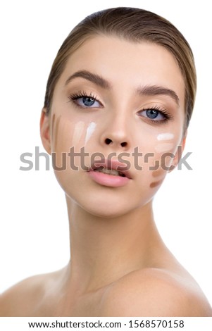 Young woman applying several  color samples of facial foundation cream at her face. Beauty model with perfect fresh skin and long eyelashes face, close up. Makeup Concept. 
