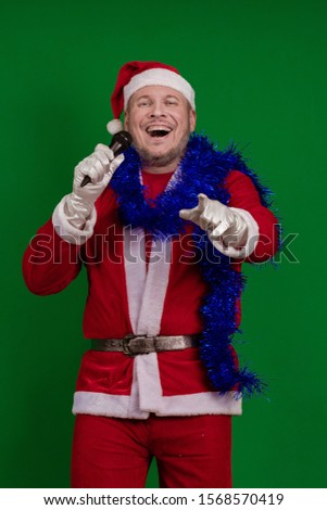 Emotional male actor in a costume of Santa Claus and a blue garland of tinsel holds a microphone in his hands, sings and gestures on a green chrome background