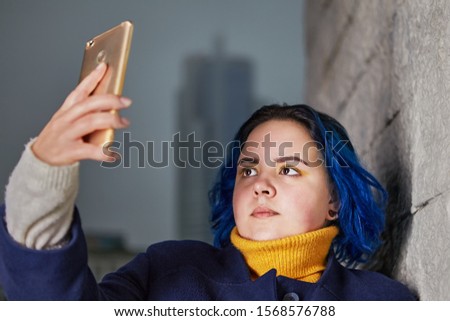 A young woman with dyed blue hair and a coat of the same color uses a smartphone to take a selfie, or to shoot video outdoors in the evening.  A chubby girl photographs herself with a mobile phone.