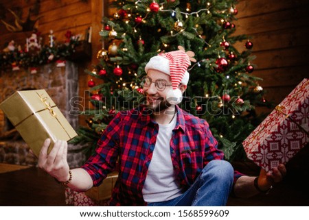 An adult man wearing glasses and a Christmas hat sitting with two presents in front of a Christmas tree on new year's eve