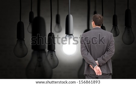 Composite image of businessman standing with hands behind back in dark room with glowing light bulb