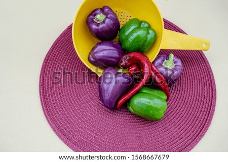 A yellow colander, red, green and purple peppers and a magenta colored placemat makes a pretty colorful picture with copy space available. Bokeh effect.