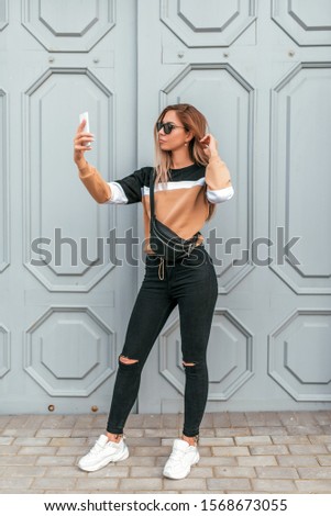 Beautiful fashionable and stylish woman summer city. Photographs himself on phone, selfie on camera, modern hipster clothes, sweaters and jeans. Handbag over shoulder. Sunglasses