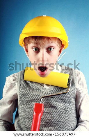 Beautiful cheerful blond boy wearing a yellow hard hat holding a painting roller looking surprised on blue background