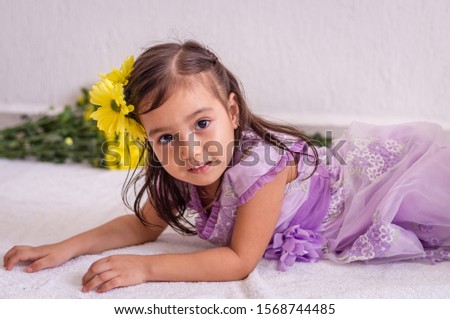 Little girl with yellow flowers in her hair, lying down, looks at camera and flowers in the background