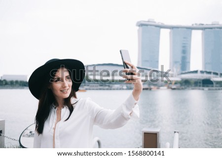 Charming touristic woman with confident smile standing at pier and taking selfie on mobile phone on background of designed architectural building and river