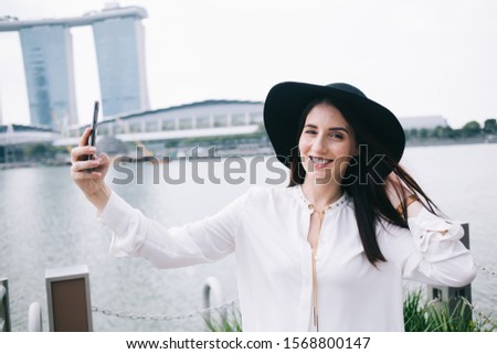 Fashion touristic woman with smile holding hat while standing at pier and taking selfie on mobile phone on background of designed architectural building and river 