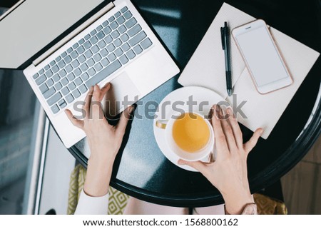 From above crop adult female in formal clothes interacting with laptop while sitting at black round table with notepad and smartphone and drinking beverage in modern office