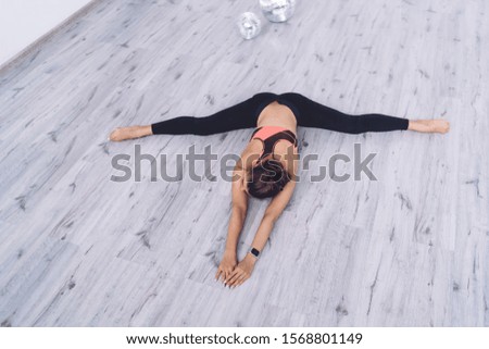From above of young brunette in black leggings doing twine stretching and leaning forward on floor training in modern studio