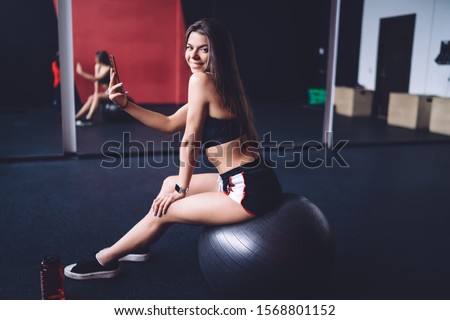 Side view of flirty smiling fit female taking selfie with mobile phone while sitting on exercise ball in gymnastic room and looking over shoulder at camera