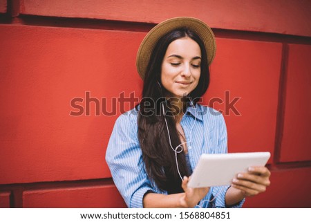 Glad young female in casual outfit and summer hat browsing tablet and enjoying music in earphones while standing near red wall in city