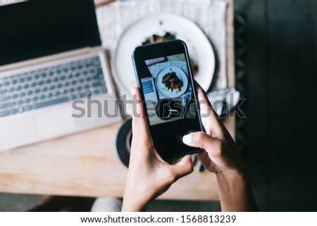 Crop hands of modern female taking picture of served dish and cup of coffee sitting at wooden table with laptop