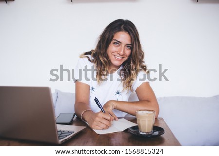 Happy cheerful young woman writing in notebook and making to do list while sitting in cafe with laptop and cup of coffee