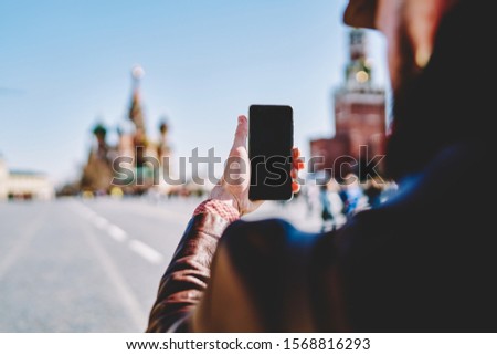 Back view of crop male holding mobile phone with blank black screen and taking photo while visiting Red Square in sunny spring day