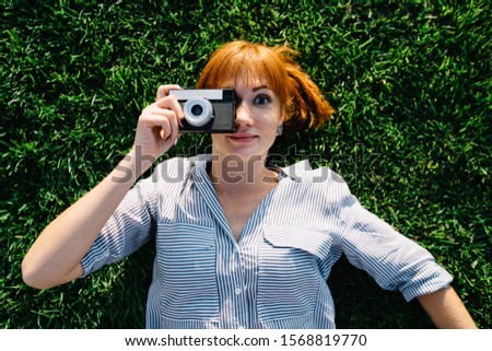 Portrait of funny caucasian woman lying on green grass making picture on vintage camera during free time,female photographing amateur satisfied with old retro equipment taking pictures outdoors