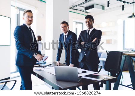 Multiracial team of serious coworkers in suits looking at camera while standing at table during working on business project in modern office room