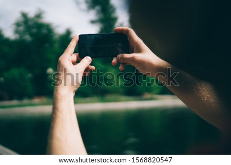 Unrecognizable man using modern smartphone to take picture of blurred pond while resting in green park on sunny summer day