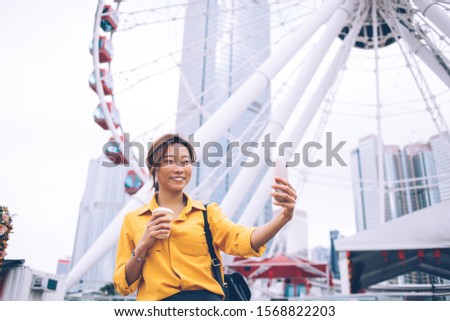 Cheerful Japanese female influencer blogger shooting video vlog during time for entertainment in Chinese metropolis Hong Kong, happy Asian woman clicking selfie near observation wheel ferris