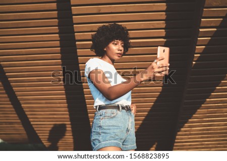Trendy dressed african american millennial female influencers in casual good look using mobile phone for making video for vlog, attractive dark skinned woman posing for selfie via app and smartphone 