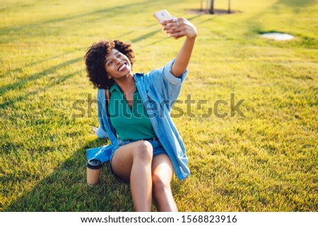 Cheerful african american hipster girl with curly hair posing on green grass in park resting on weekends, happy dark skinned woman using modern camera on cellular for making video for vlog