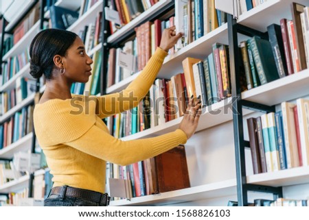 Low angle of smart African American female in casual outfit taking textbook from shelf while performing research in university library