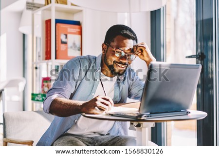 Smiling tired black guy watching on laptop and leaning with head on hand while taking notes on notebook during study in light room Royalty-Free Stock Photo #1568830126