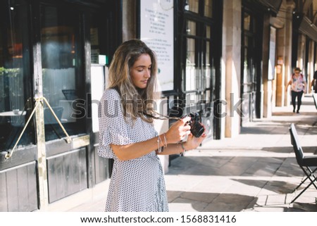 Attractive female photographer with modern camera in hand checking photos during leisure in city, beautiful woman dressed in stylish sundress watching video on technology for taking pictures
