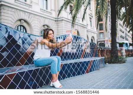 Positive joyful hipster girl showing tongue while shooting video for social networks resting on ceramic bench with smartphone device in hands, beautiful woman in casual wear taking pictures via app