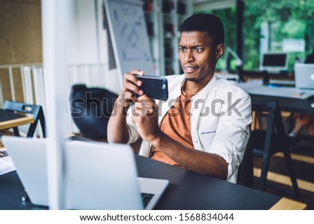 African American male student puzzled with web information on university website photographing laptop screen for sending media sms to colleagues and discuss mistakes, user of modern technology