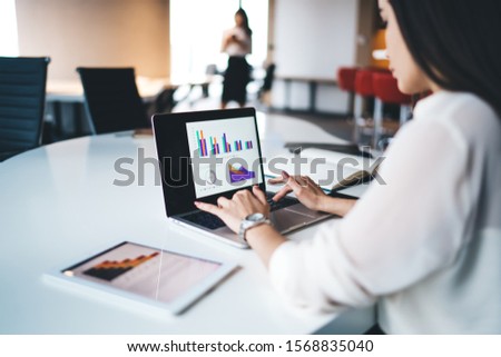 Blurred elegant young lady browsing laptop and analyzing schemes and data while sitting at table in modern office and working