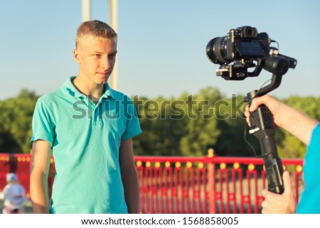 Boy teenager model and male photographer taking photos videos with camera and stabilizer, filming backstage, background foot bridge over the river on sunny summer day