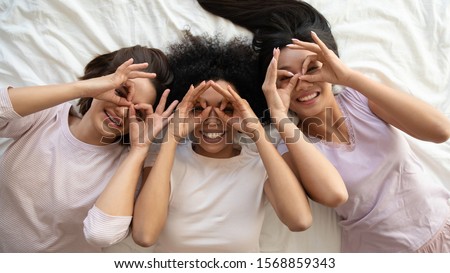 Funny pretty multiracial girls wear sleepwear having fun look at camera relax on bed, three diverse women friends laugh lying on bed together celebrate sleepover pajama hen party, top view portrait