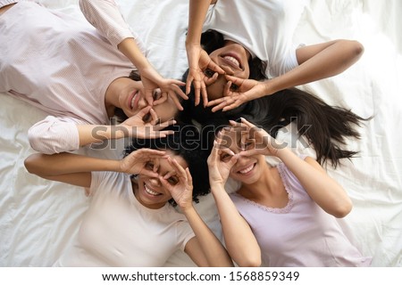 Four smiling young multi ethnic ladies lying on bed look at camera make binoculars, happy multiracial girls friends wear pyjamas having fun laugh together enjoy pajama hen party, top view portrait