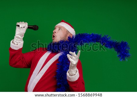 Emotional male actor in a costume of Santa Claus and a blue garland of tinsel holds a microphone in his hands, sings and gestures on a green chrome background