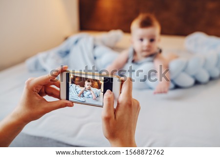 Mother taking photo of her adorable baby boy. Baby lying on stomach on bed in bedroom.