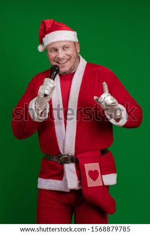 Male actor in a costume of Santa Claus holds a microphone in his hands, sings and poses on a green background