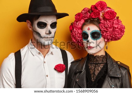 Calm young woman and man wears skull makeup, female in beautiful floral wreath, dressed in halloween holiday costumes, keep eyes shut, isolated over yellow studio background. People, Day of Dead