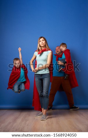 Smiling mom and dad, children superheroes in red cloaks on blank blue background