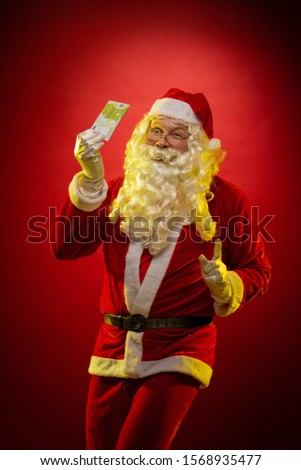 Male actor in a costume of Santa Claus holds in his hands banknotes, money and posing on a dark red background
