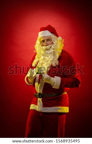 Male actor in a costume of Santa Claus holds in his hands banknotes, money and posing on a dark red background