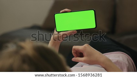 Young woman lying on a couch and using smartphone with horizontal green screen