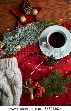 A cup of coffee and a garland on a wooden tray with a cozy red sweater and a warm hat. New Year's still life with a fir branch and mountain ash