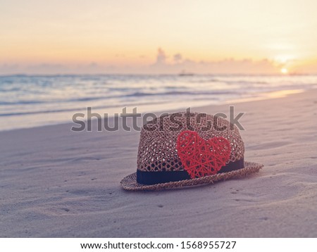 A straw hat and a red wicker heart are on the sandy beach. The sea or the ocean and the sky with the rising sun are in the background. Tropical, exotic, beach valentine's day template.