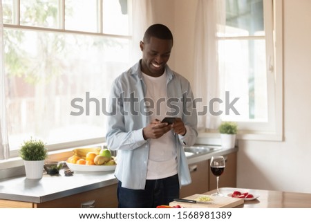 Smiling african American man use smartphone texting messaging preparing for romantic date at home, happy biracial male cellphone user relax on leisure weekend cooking in modern own kitchen